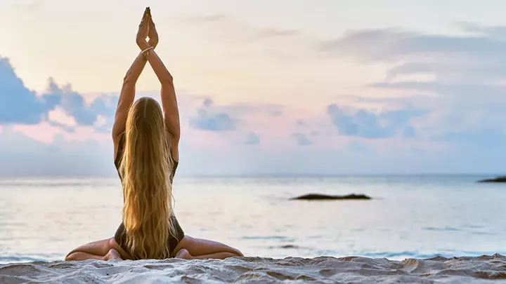 A young woman at the beach, doing yoga in a kneeling position with hands towards the sky and palms together, her back is turned to us with long hair flowing down her back